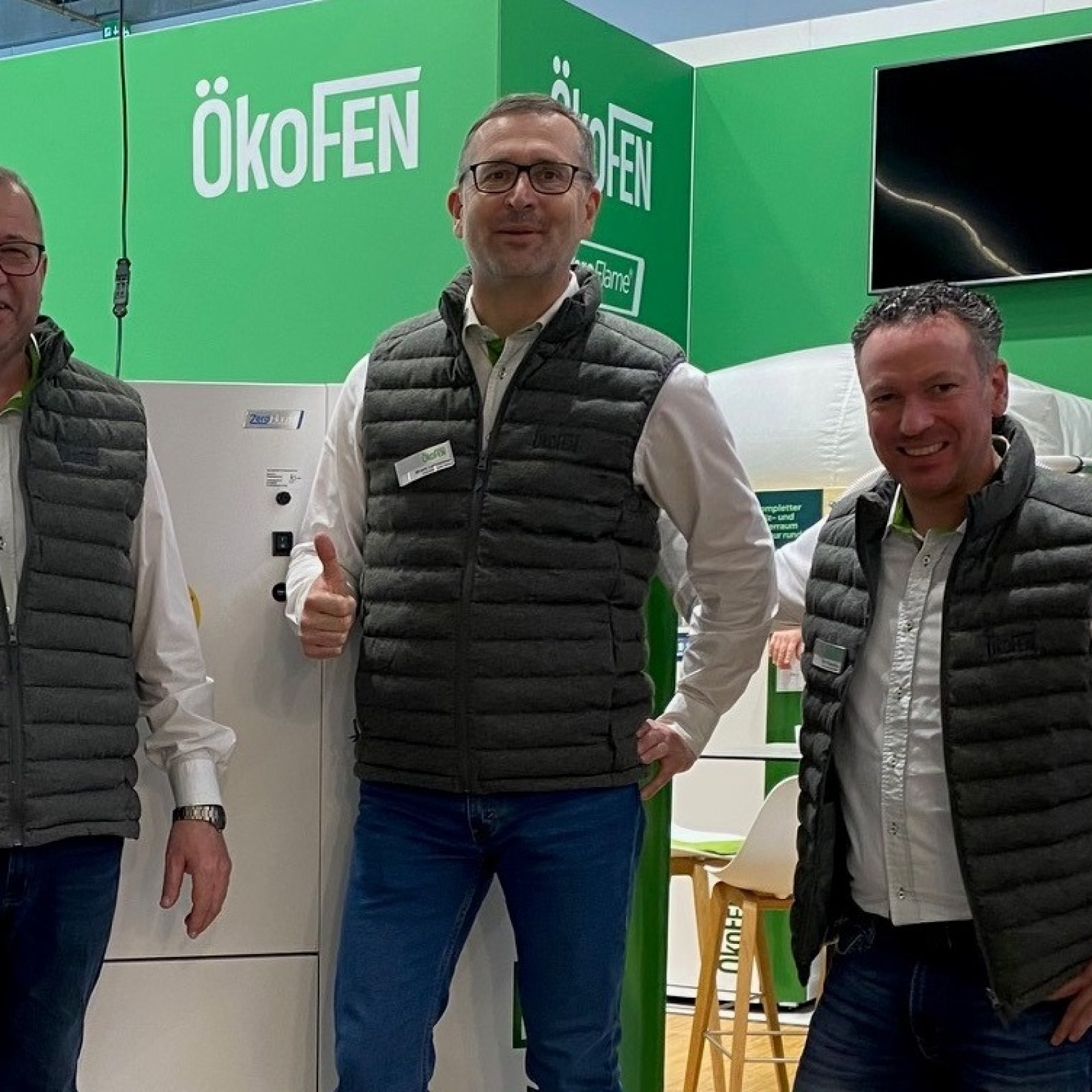 Vier Männer in Westen vor ÖkoFEN-Stand auf einer Messe, lächelnd in die Kamera schauend.